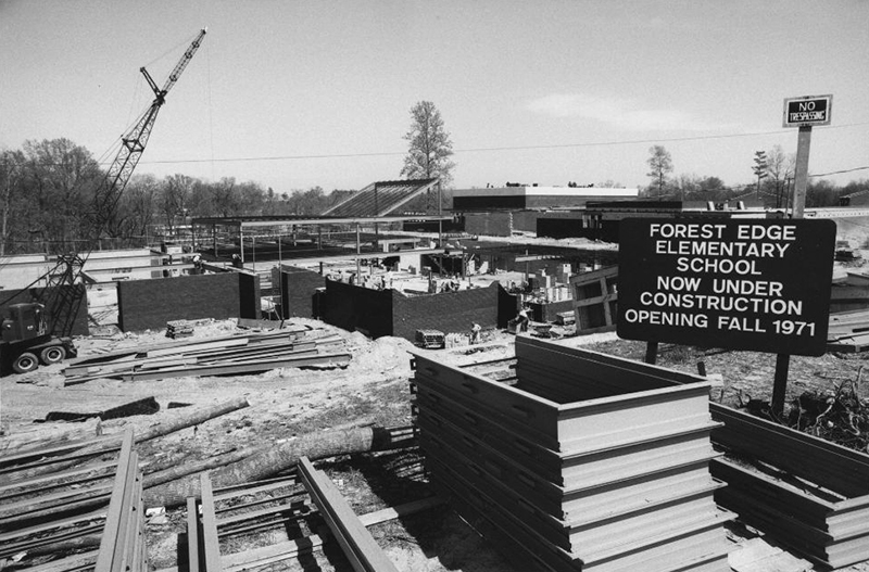 Black and white photograph of the construction of Forest Edge Elementary School. In the foreground, there is a sign announcing the name of the school and that it will be opening in fall of 1971. Workmen can be seen performing various tasks. In the background, there are trees without foliage.