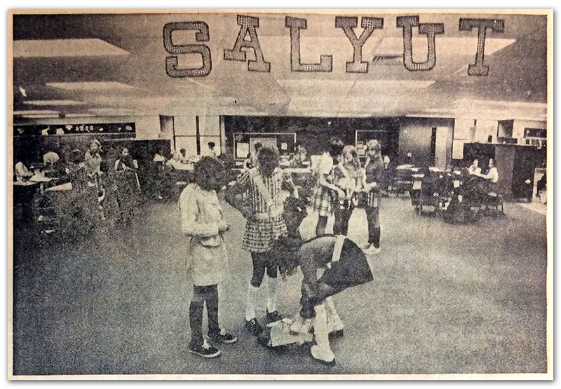 Black and white photograph of Salyut Crater that appeared in the Sun Newspaper. The clipping does not show the newspaper’s printing date, but it was likely between 1971 and 1973 because no classroom partitions are visible in the photograph. The first partitions to the learning area were added in 1973. Three safety patrol students can be seen in the foreground putting on their belts. In the background students are clustered around groups of tables. 