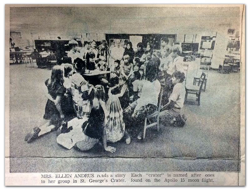 Black and white photograph of St. George’s Crater that appeared in the Sun Newspaper. The clipping does not show the newspaper’s printing date, but it was likely between 1971 and 1973 because no classroom partitions are visible in the photograph. A group of approximately 30 students are clustered around a table in the center of the crater where the teacher is reading a story. The image caption reads: Mrs. Ellen Andrus reads a story to her group in St. George’s Crater. Each crater was named after ones found on the moon by the crew of NASA’s Apollo 15 spaceflight.  