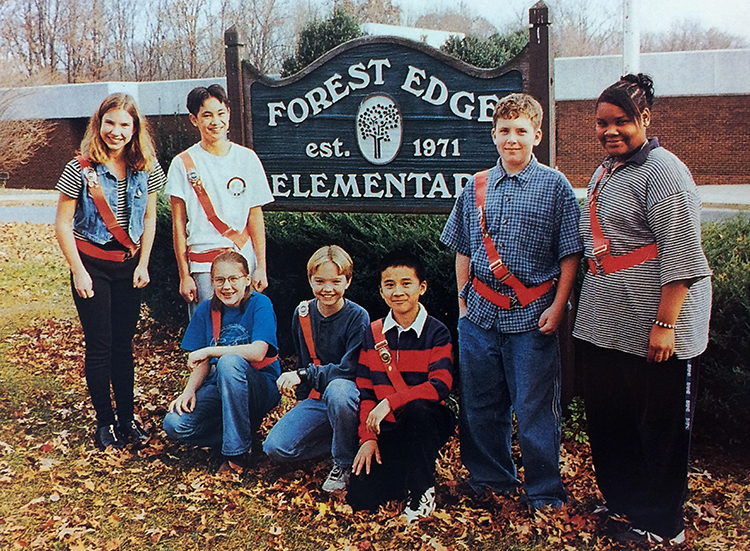 Color yearbook photograph of students who served in the Forest Edge Safety Patrol. Seven students, wearing bright orange belts, are pictured. Some are standing, some kneeling, around the sign in front of our school. The sign reads: Forest Edge Elementary, established 1971. The sign is made out of wood and is painted brown and dark green with white lettering. An emblem of a tree is in the center.  